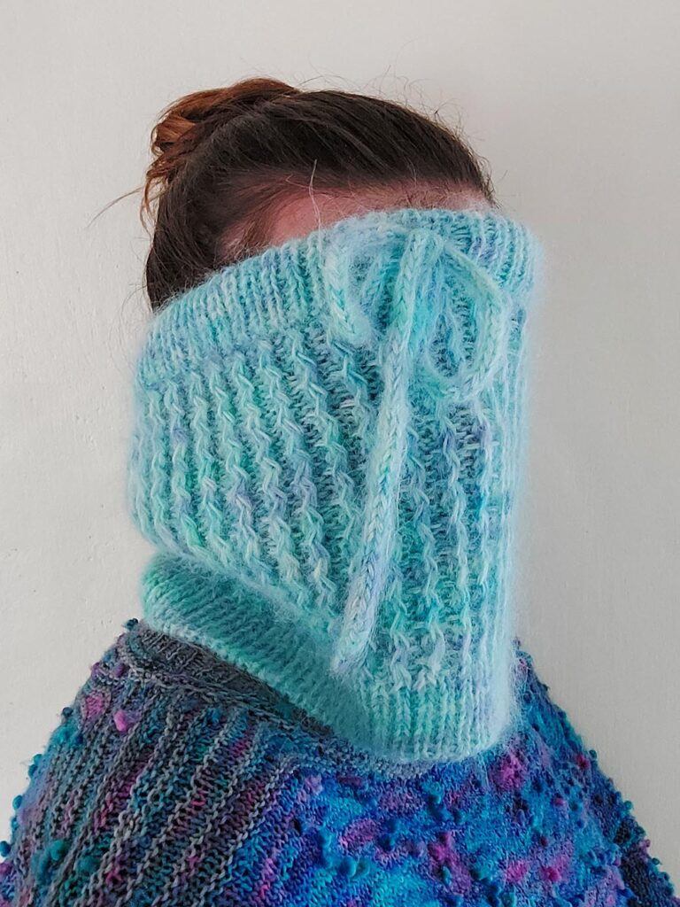 Quinzhee Cowl pulled up oven the face of the woman wearing it. Pulled up around her mouth, the I-cord tie is visible at the front with the 'zigzag' cable pattern down the body of the cowl. It has a ribbed bottom hem finished with a tubular bind off