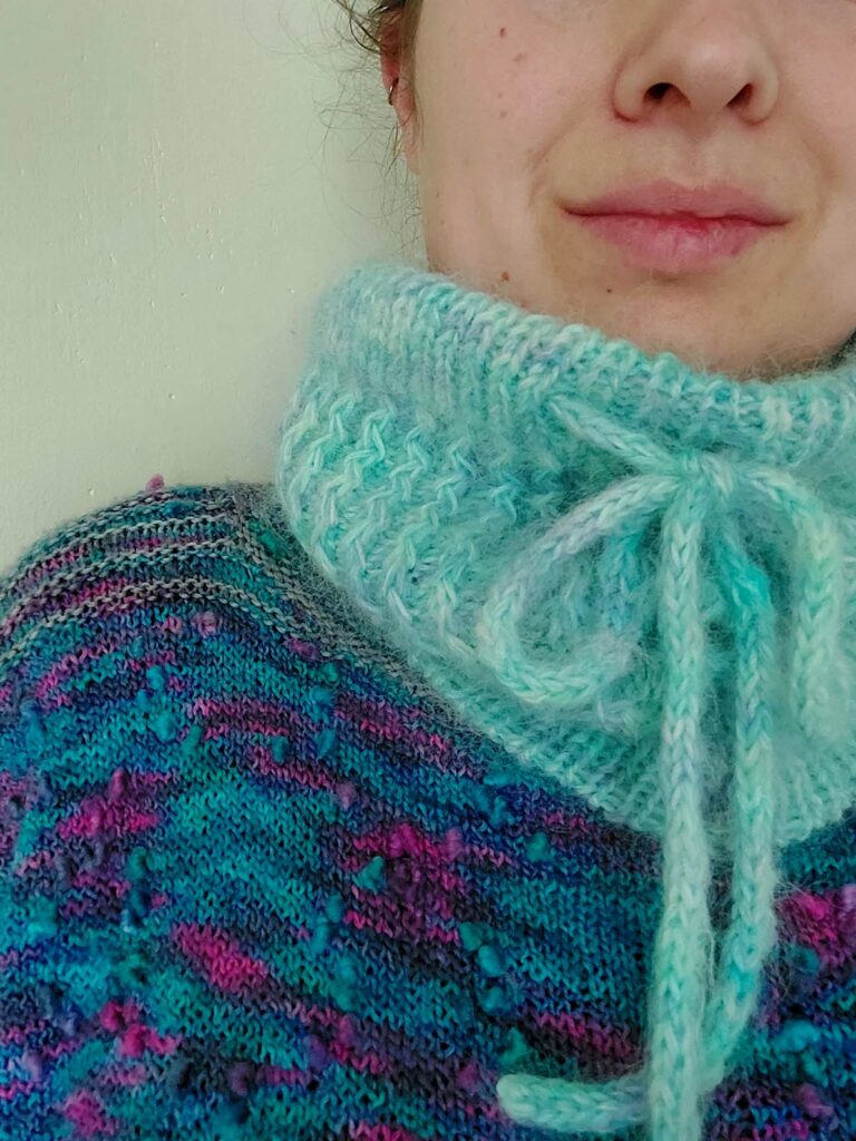 Quinzhee Cowl. Pulled up around her mouth, the I-cord tie is visible at the front with the 'zigzag' cable pattern down the body of the cowl. It has a ribbed bottom hem finished with a tubular bind off