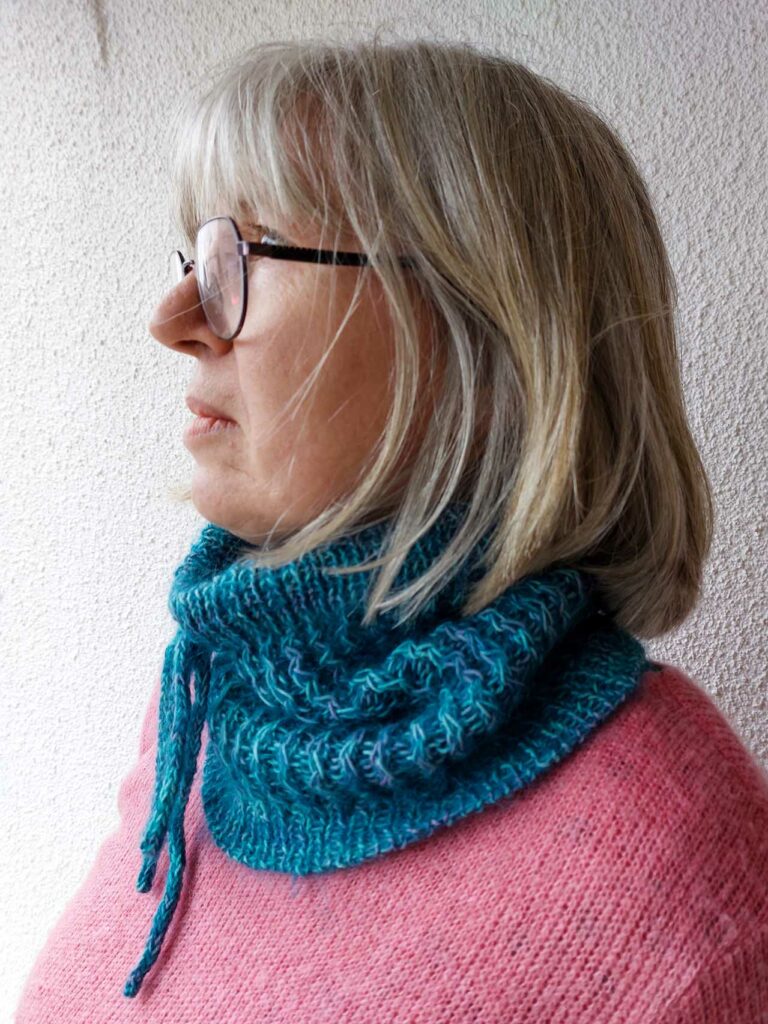 Quinzhee Cowl. Pulled up around her mouth, the I-cord tie is visible at the front with the 'zigzag' cable pattern down the body of the cowl. It has a ribbed bottom hem finished with a tubular bind off