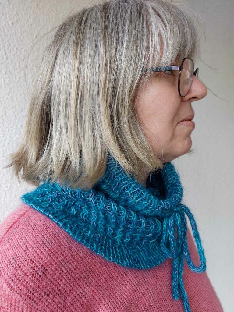 Quinzhee Cowl. Pulled up around her mouth, the I-cord tie is visible at the front with the 'zigzag' cable pattern down the body of the cowl. It has a ribbed bottom hem finished with a tubular bind off