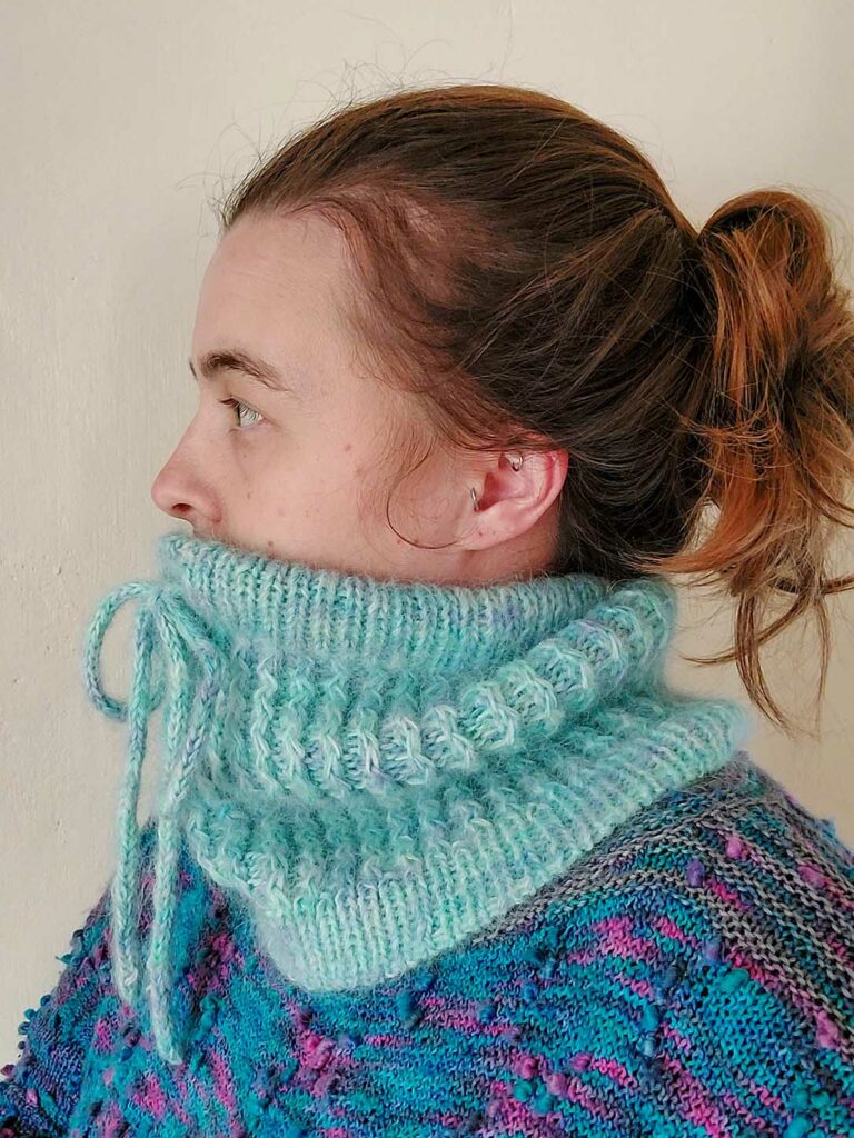 side view of a woman wearing the Quinzhee Cowl. Pulled up around her mouth, the I-cord tie is visible at the front with the 'zigzag' cable pattern down the body of the cowl. It has a ribbed bottom hem finished with a tubular bind off