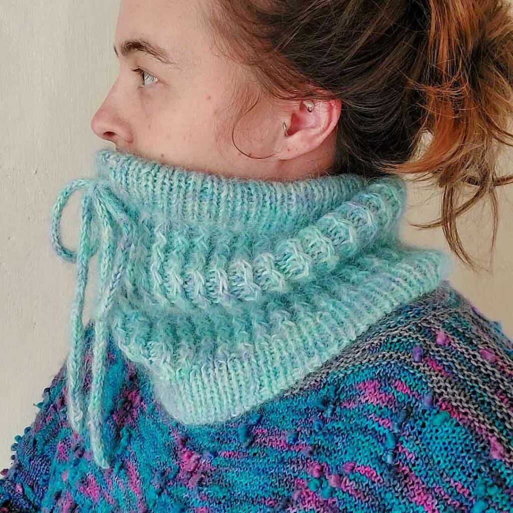 side view of a woman wearing the Quinzhee Cowl. Pulled up around her mouth, the I-cord tie is visible at the front with the 'zigzag' cable pattern down the body of the cowl. It has a ribbed bottom hem finished with a tubular bind off