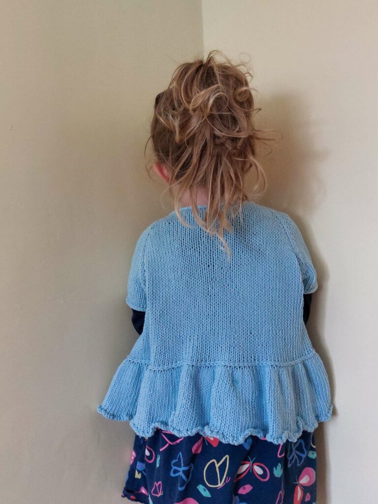 back view of the flutterbye cardigan blue cotton short sleeved version. the cardigan has an i-cord neckline, integrated icord button bands and a frilled peplum bottom, separated from the main body by a lateral (vikkel/estonian) braid.