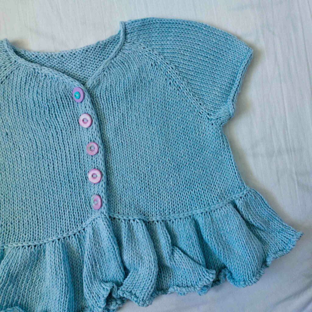 Flutterbye cardigan blue cotton short sleeve version flatlay photo The cardigan has an i-cord neckline, integrated icord button bands and a frilled peplum bottom, separated from the main body by a lateral (vikkel/estonian) braid