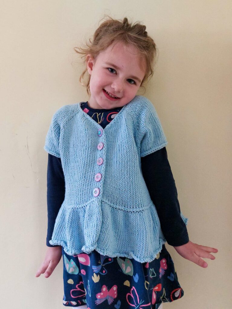 front view of the flutterbye cardigan knit in blue cotton yarn. the cardigan has an i-cord neckline, integrated icord button bands and a frilled peplum bottom, separated from the main body by a lateral (vikkel/estonian) braid