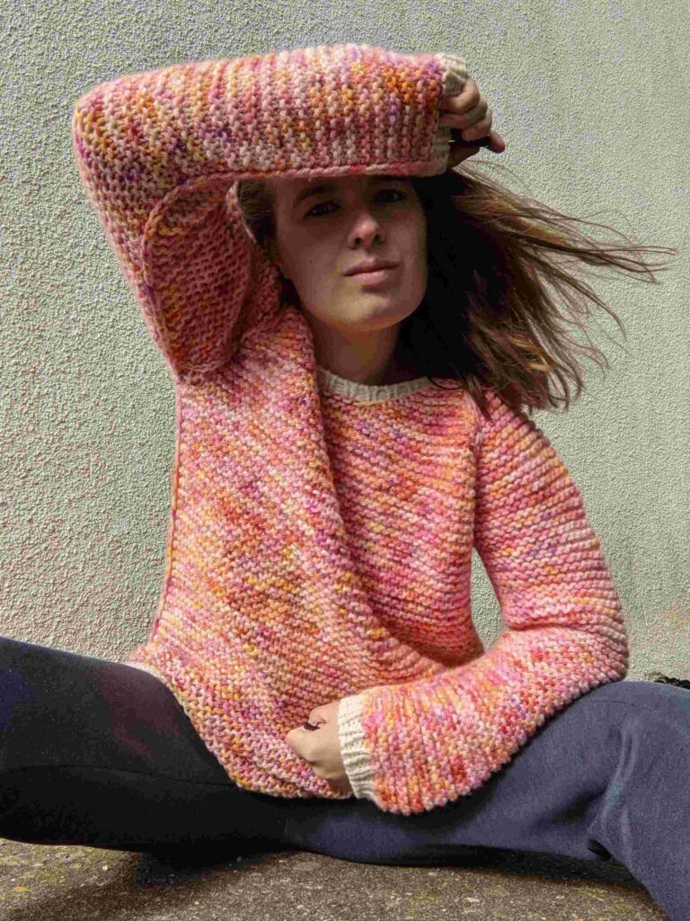 woman wearing the garter squish adult jumper with an arm over her head. showing the all over garter stitch pattern, body side slipped knit stitches which also run along the arm from under the armpit