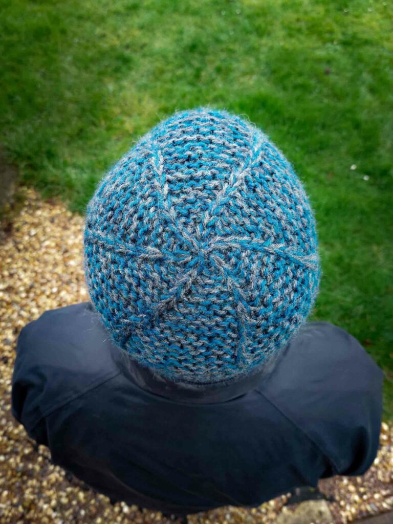 top down view of the head and shoulders of a man wearing a black coat and knitted beanie on his head. The beanie is a blue and grey marled knitted fabric in garter stitch and a swirl is created by increasing from the centre crown