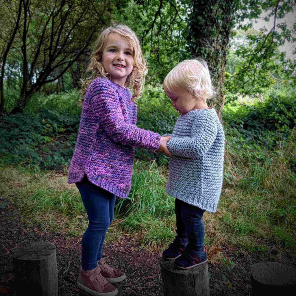 two children wearing the garter squish kids jumper - side view. on show is the all over garter stitch pattern, slip knit stitch detail running down the side of the body and dropped back hem on the boy and the dropped front and back hem on the girl