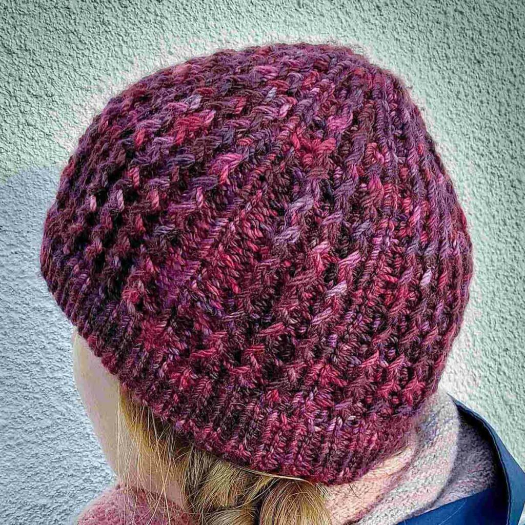 back view of the quinzhee beanie Knit from the top down, the quinzhee beanie features small zigzag like cables which make the hat very warm as squishy to wear. This beanie has a folded secured brim and is knit from a marron/wine coloured variegated hand dyed yarn