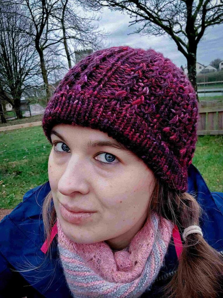 woman wearing the quinzhee beanie, smiling at the camera. Knit from the top down, the quinzhee beanie features small zigzag like cables which make the hat very warm as squishy to wear. This beanie has a folded secured brim and is knit from a marron/wine coloured variegated hand dyed yarn