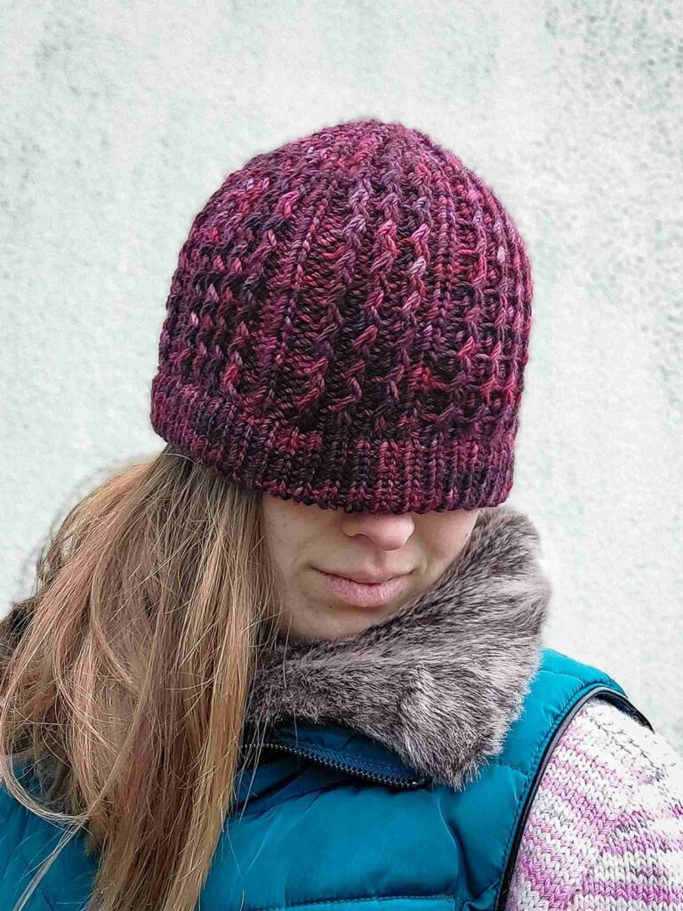 woman wearing the quinzhee beanie, looking down to show the cable pattern. Knit from the top down, the quinzhee beanie features small zigzag like cables which make the hat very warm as squishy to wear. This beanie has a folded secured brim and is knit from a marron/wine coloured variegated hand dyed yarn