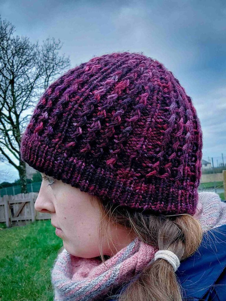 side view of the quinzhee beanie. Knit from the top down, the quinzhee beanie features small zigzag like cables which make the hat very warm as squishy to wear. This beanie has a folded secured brim and is knit from a marron/wine coloured variegated hand dyed yarn